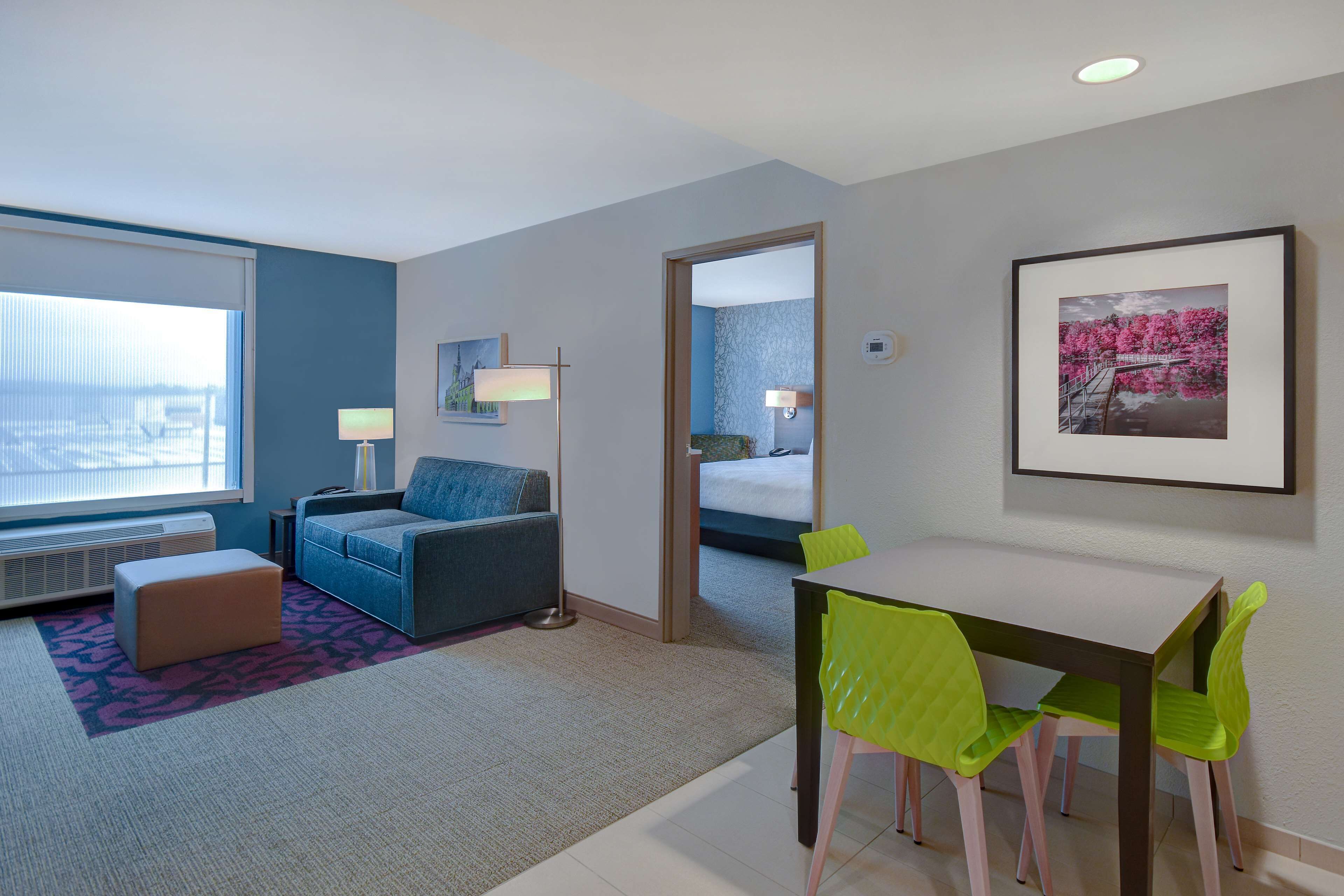 A modern hotel room with a large window, a sofa, a dining table with chairs, and a framed picture on the wall.