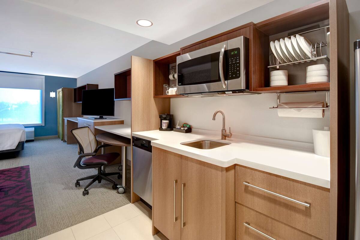 A modern kitchen with a microwave, sink, and cabinets. There is a desk with a chair and a television in the background.