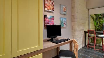 A room with a desk, computer, and chair, a window with a plant, and artwork on the wall.