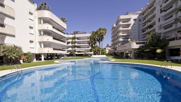A swimming pool surrounded by multi-story buildings with balconies, palm trees, and a clear blue sky.