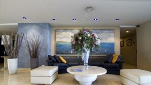 A modern lobby with a round marble coffee table, two white sofas, a large painting of a seascape, and tall decorative plants.