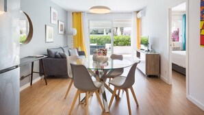 A modern living room with a glass dining table, grey sofas, and a large window with yellow curtains.