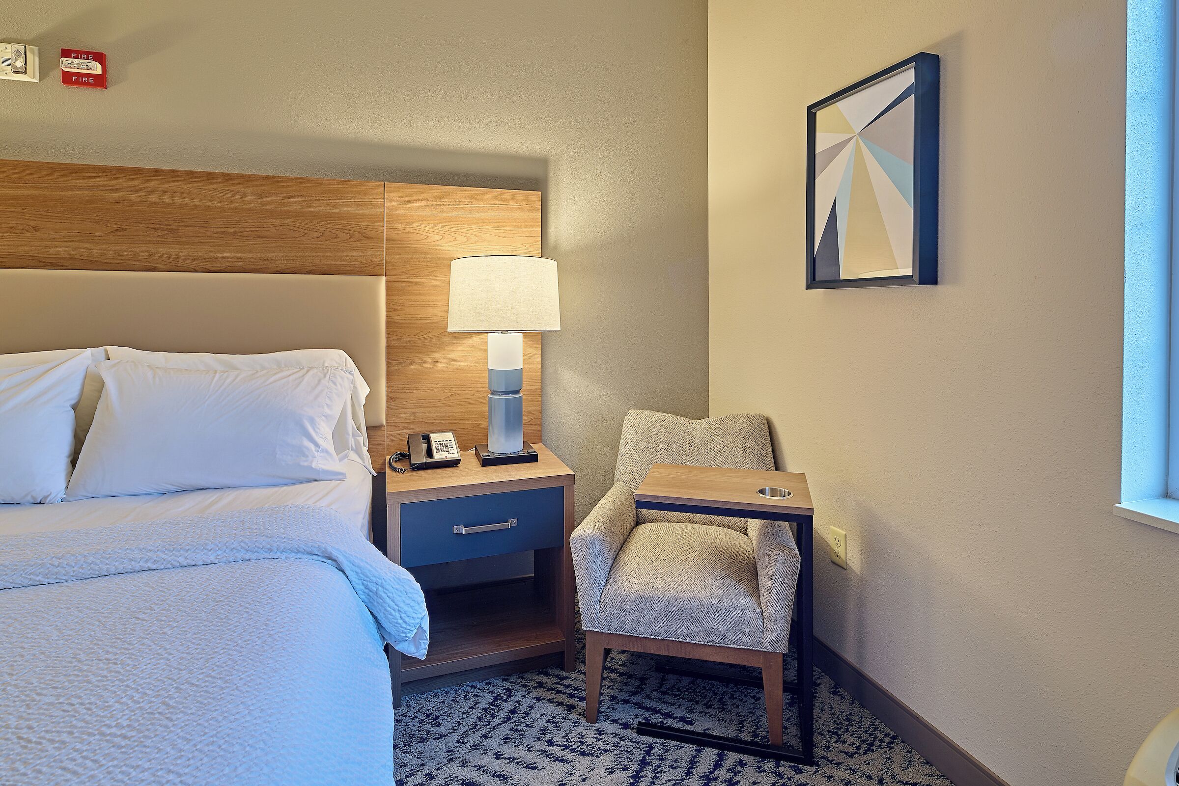 Une chambre d’hôtel comprenant un lit, un bureau avec une lampe, une chaise, une table de chevet et un tableau accroché au mur.