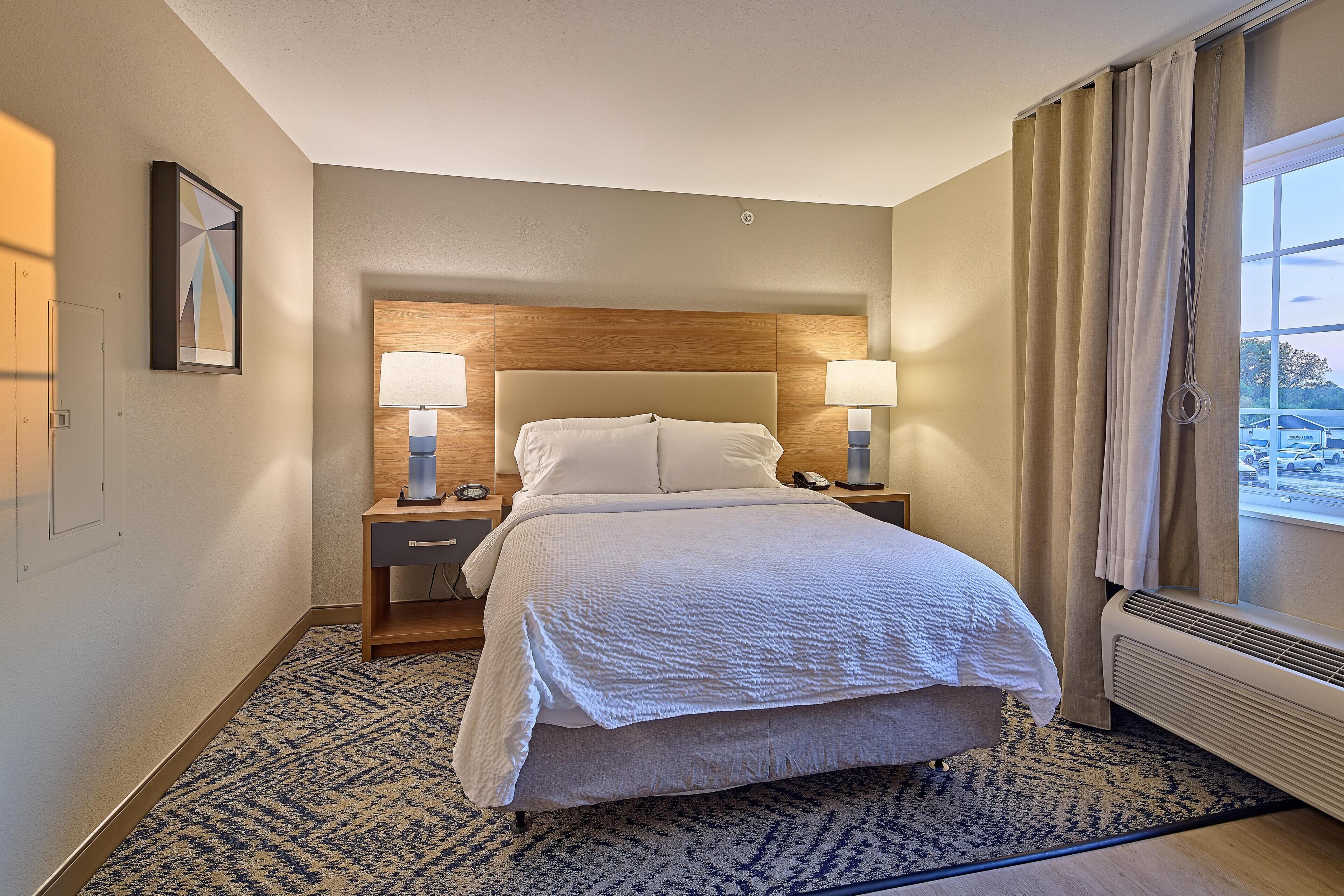 Une chambre d’hôtel comprenant un lit, des lampes de chevet, une table de nuit, une fenêtre avec des rideaux et un climatiseur fixé au mur.