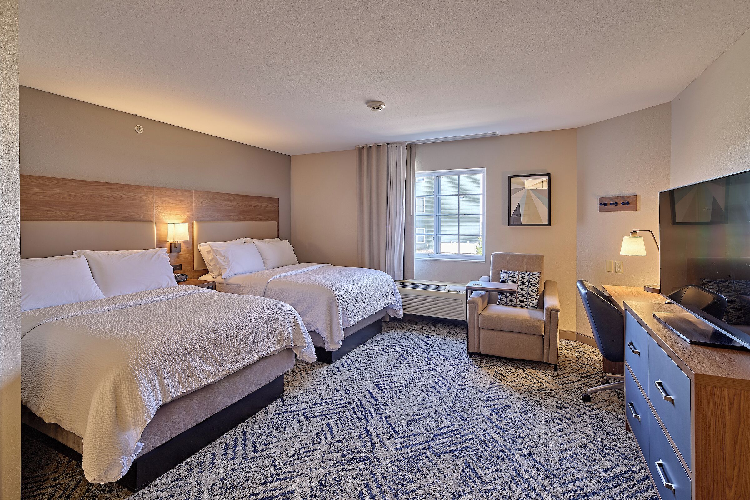 Une chambre d’hôtel avec deux lits, un bureau, une chaise, une télévision et une grande fenêtre.
