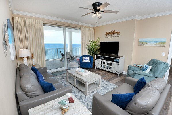 A living room with a sofa, armchairs, a coffee table, a TV, and a balcony with a view of the ocean.