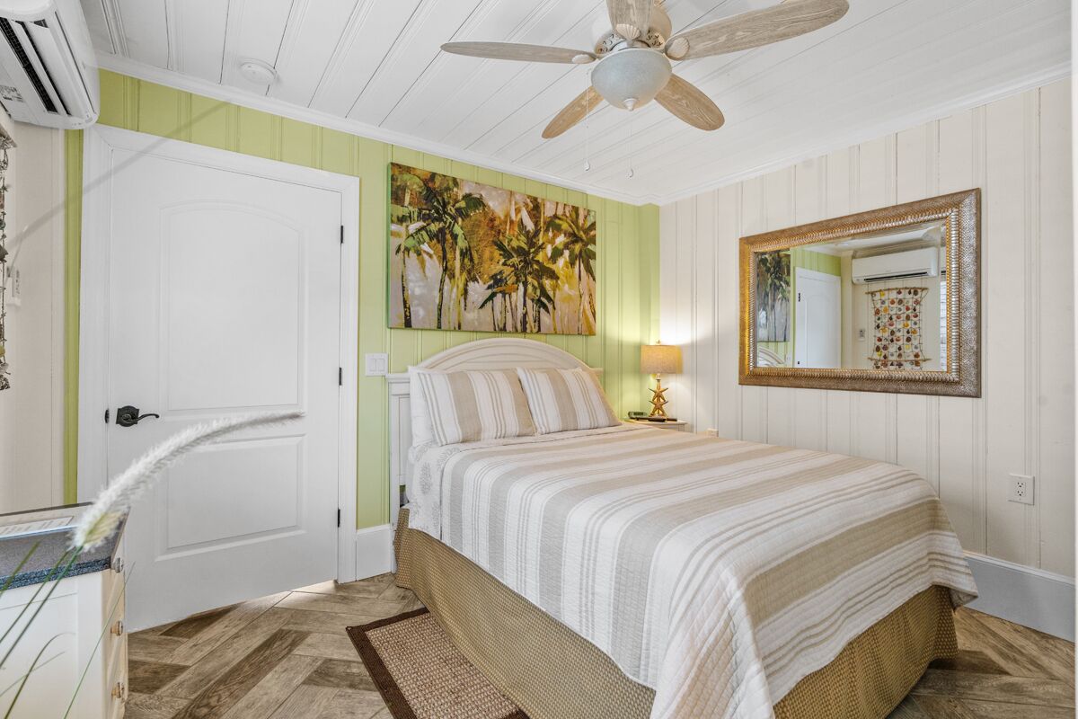 A bedroom with a bed, a ceiling fan, a painting, a mirror, and a door.