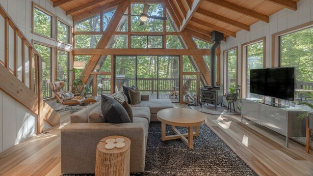 Ein geräumiges Wohnzimmer mit einem großen Sofa, einem Couchtisch und einem Fernseher. Große Fenster und eine Holzbalkendecke sind vorhanden.