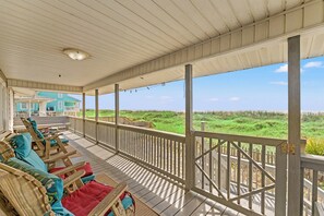 Un porche cubierto con muebles de madera, vista a la playa y una casa al fondo.