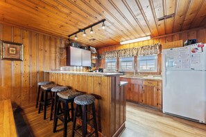 Interior de una cabaña de madera con barra, alacenas de madera y un refrigerador.