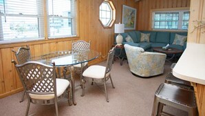 A room with a round glass table and metal chairs, a teal sofa, and a patterned armchair. - Nautical Winds, Beach House in Cherry Grove, Just Steps From the Beach (North Myrtle Beach)