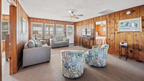 Un salon spacieux avec un mur lambrissé en bois, un ventilateur de plafond, un canapé gris, deux fauteuils à motifs et une petite table.