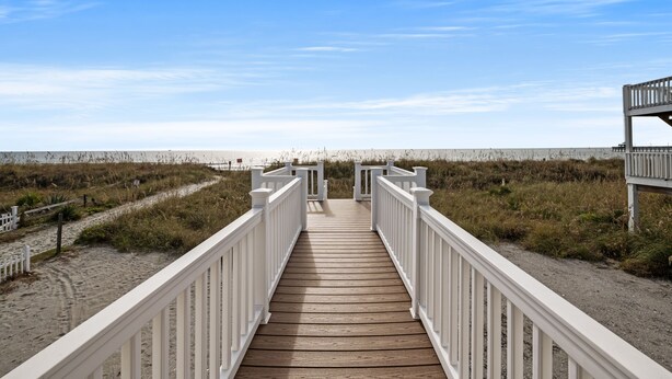 Une promenade en bois menant à une plage, avec des garde-corps blancs et un sentier sablonneux.