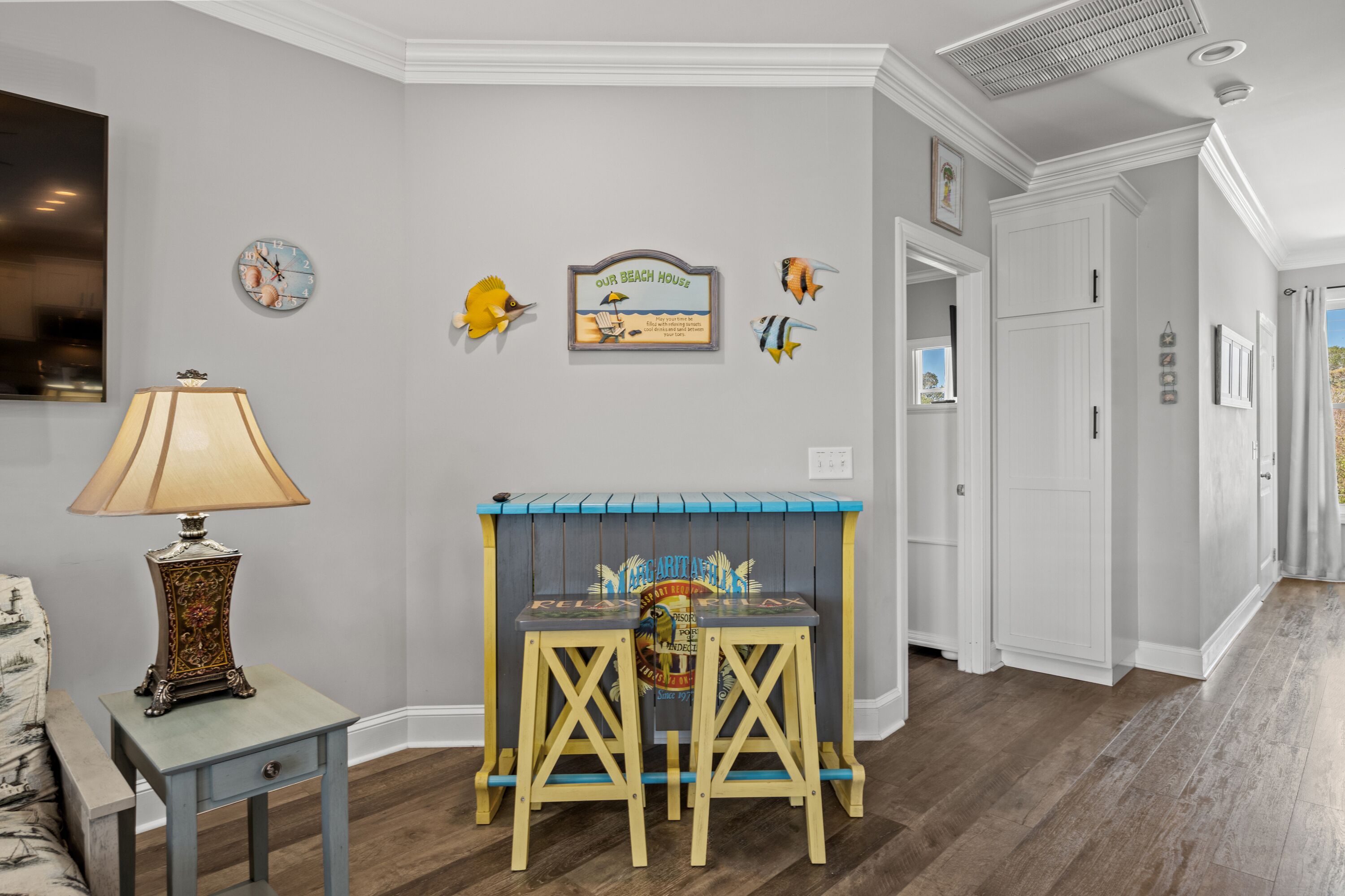 A hallway with a wooden floor, a small table with a lamp, and wall decorations including a clock and a sign.