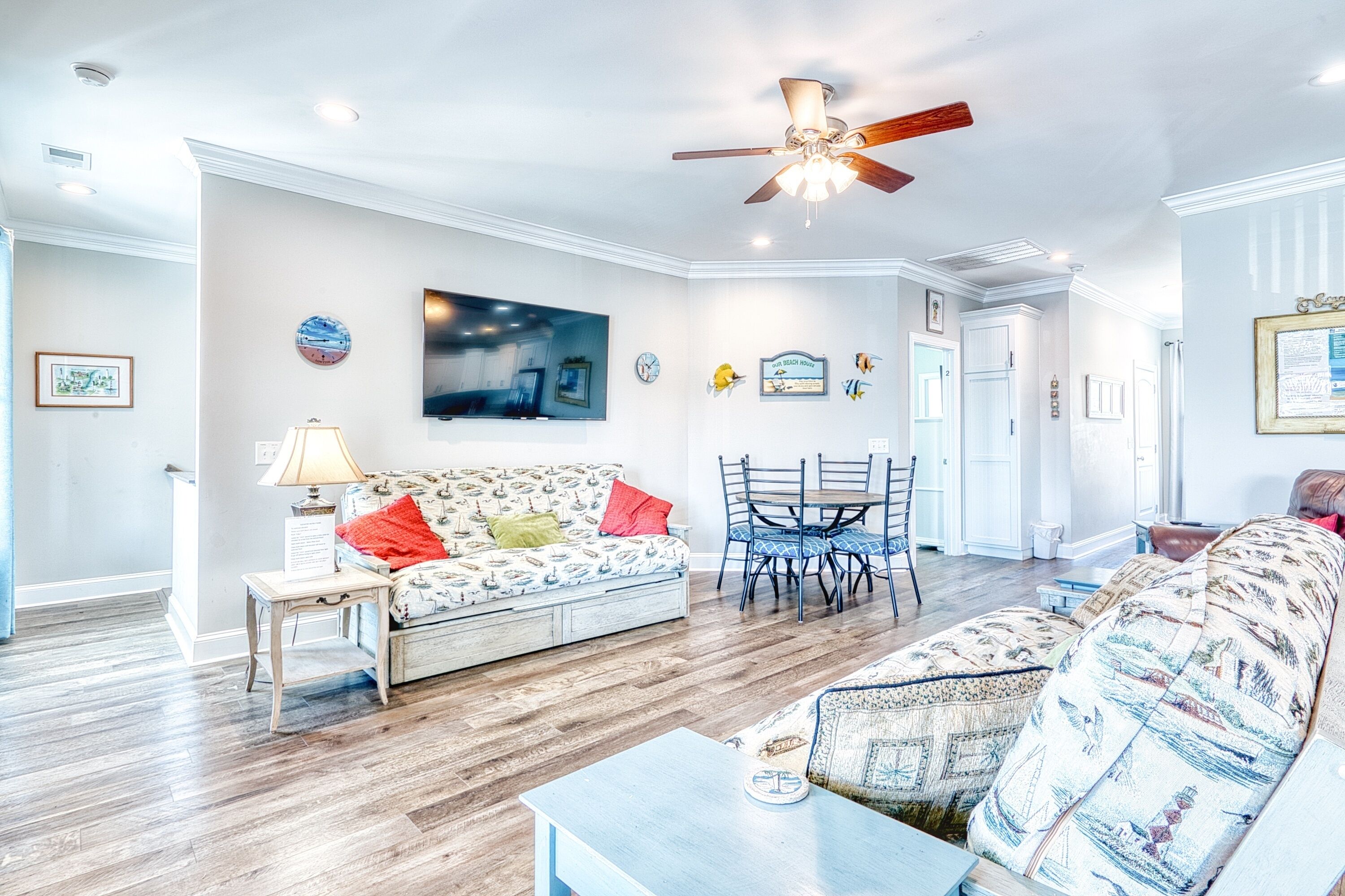 A spacious living room with a sofa, a coffee table, a dining table with chairs, a ceiling fan, and a flat-screen TV mounted on the wall.
