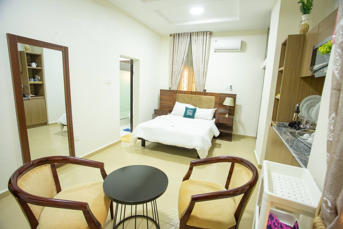 A hotel room with a bed, two chairs, a small table, a mirror, a wardrobe, and a kitchen area.