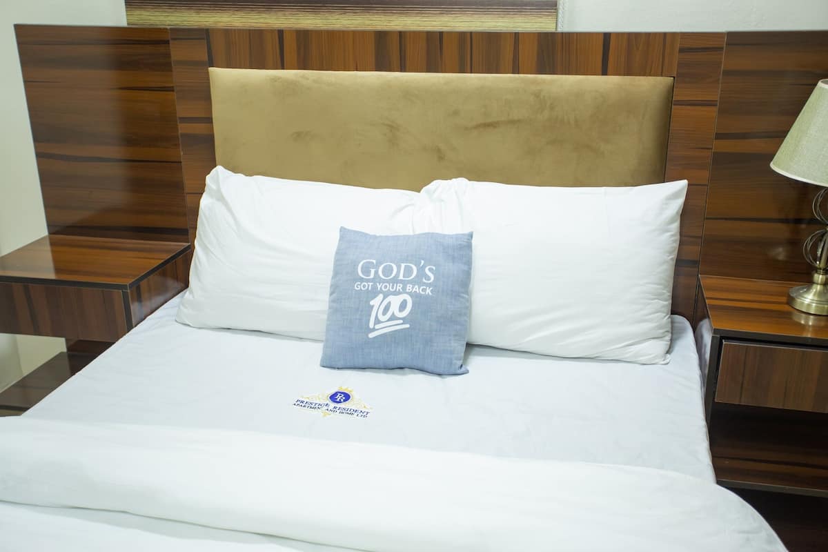 A neatly made bed with a pillow that reads "GOD'S GOT YOUR BACK 100" and a logo on the bedspread.