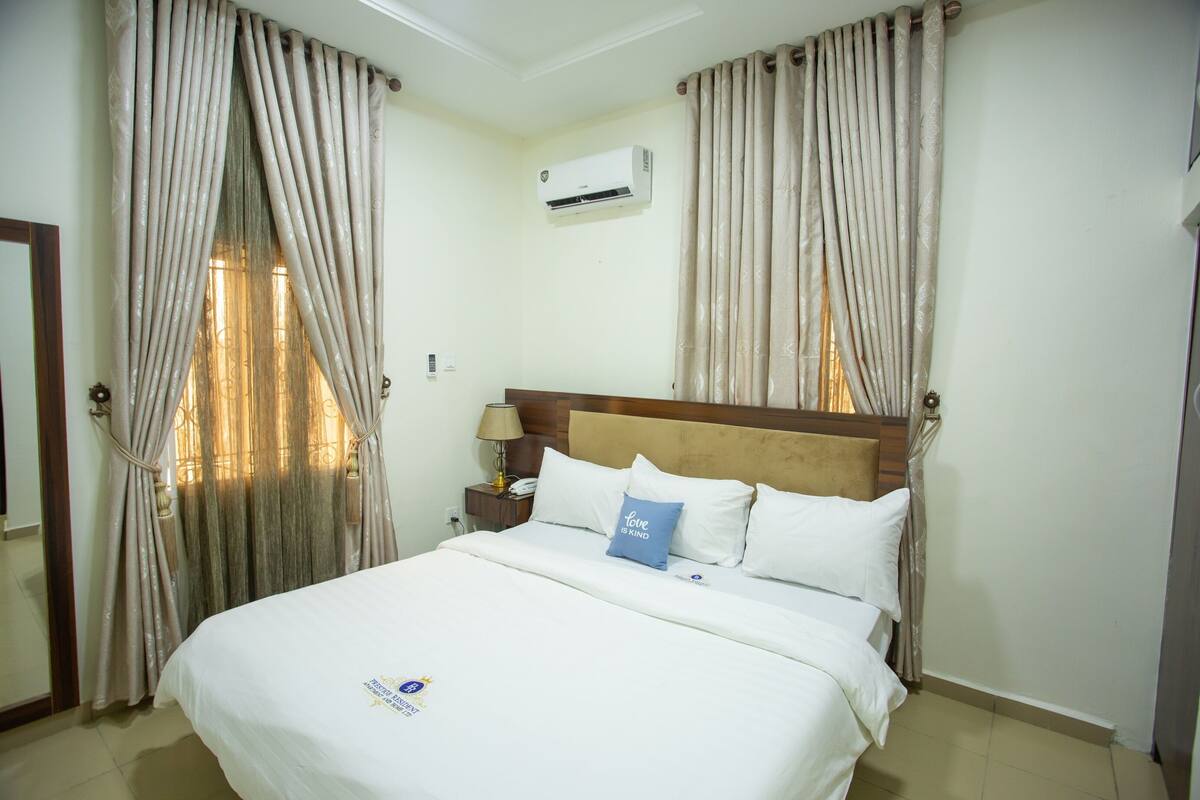 A hotel room with a bed, curtains, an air conditioning unit, a nightstand, and a lamp.