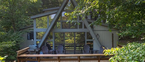 A wooden deck with Adirondack chairs and a grill, overlooking a forested area.