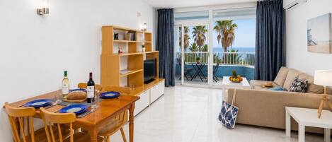 Un appartement moderne comprenant un coin repas, un salon avec un canapé et une télévision, un balcon offrant une vue sur les palmiers et la mer, ainsi qu’une grande fenêtre avec des rideaux.
