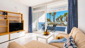 A modern living room with a television, a coffee table with fruit, a sofa, and a balcony with a view of palm trees. - Jardines Apartment 1 (Puerto del Carmen)