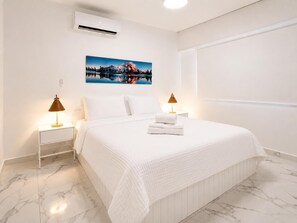 Habitación de hotel con cama, paredes blancas, un cuadro, dos lámparas de noche y suelo de mármol.