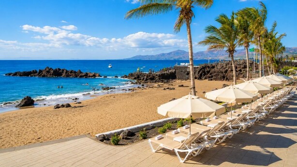 A beach with lounge chairs and umbrellas, a rocky shoreline, and a clear blue sky. - Jardines Apartment 1 (Puerto del Carmen)