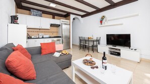 Una sala de estar moderna con un sofá gris, cojines rojos, una mesa de centro blanca con una botella y vasos, un televisor sobre un mueble blanco, y una cocina al fondo.