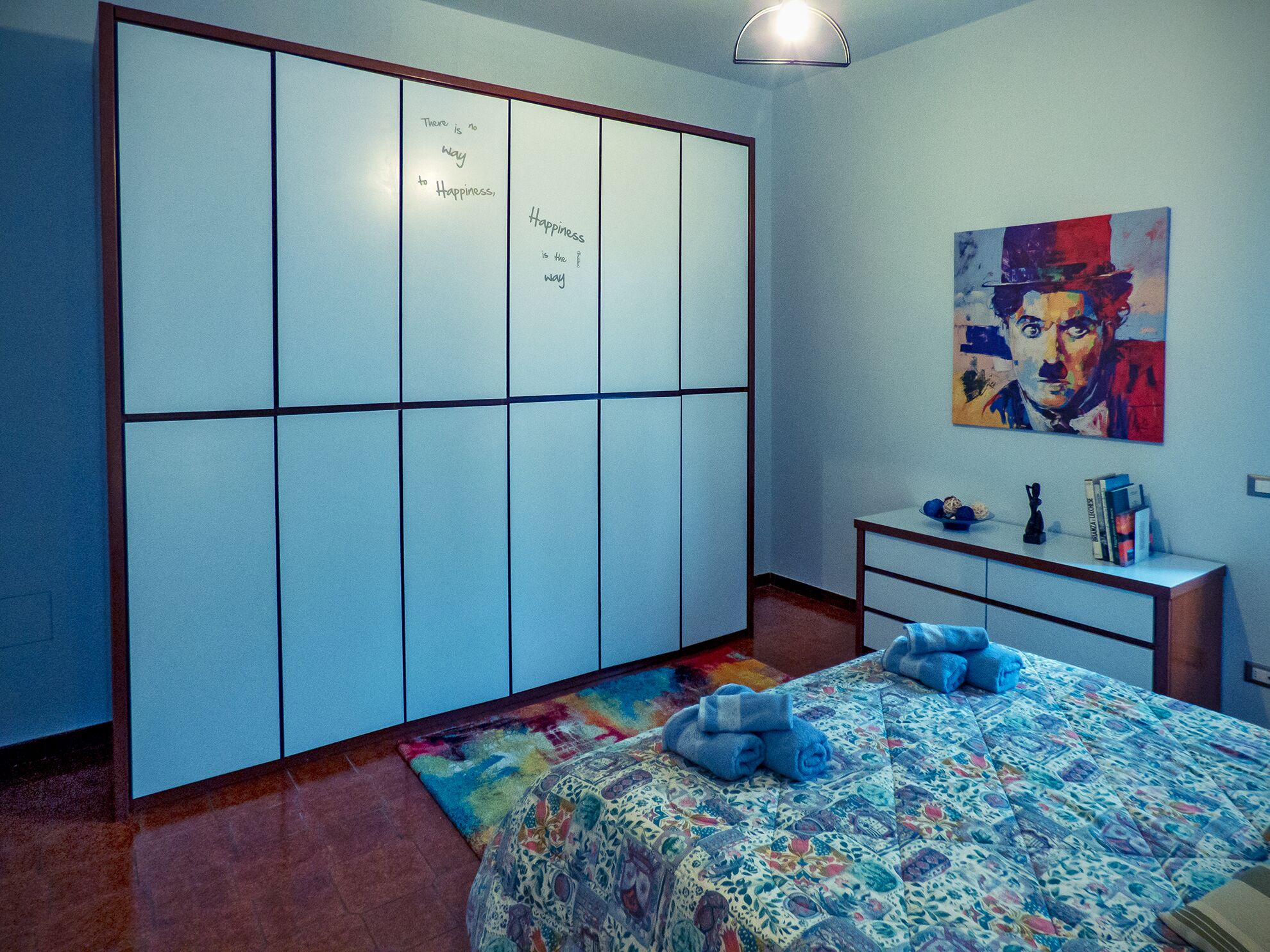 A bedroom with a bed, a wardrobe, a desk, and a colorful painting on the wall.