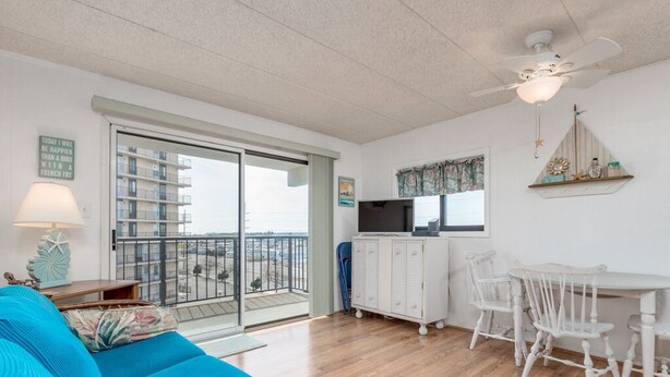 Un salon lumineux avec un canapé bleu, une table à manger avec des chaises, un ventilateur de plafond et une vue sur un balcon doté d’une balustrade.