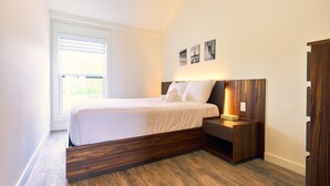 A modern bedroom with a wooden headboard, a bedside table with a lamp, and a window with blinds. - Cottage 18: Lower-Level Beachfront Unit in 4-Plex (Wainfleet)