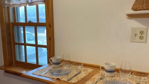 Un comedor con una mesa de madera dispuesta para dos, una ventana con vista a la calle y un estante con una cesta.