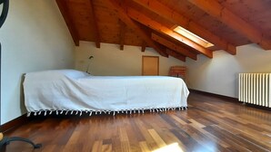 Une pièce avec un plancher en bois, un lit recouvert d’une courtepointe blanche, un radiateur et une lucarne.