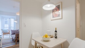 Ein Essbereich mit einem weißen Tisch, Stühlen, einer Obstschüssel, einer Weinflasche und einem gerahmten Bild an der Wand.