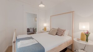 Ein ordentlich bezogenes Bett mit weißen Bettwäsche, einer blauen Zudecke und einem hölzernen Kopfteil. Ein kleiner Nachttisch mit Lampe und Blumenvase steht daneben. Die Wände sind weiß und der Boden ist aus Holz.