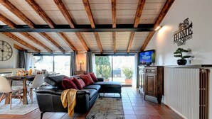 Una sala de estar con un sofá negro, vigas de madera, una mesa de comedor con sillas, un televisor y un radiador.