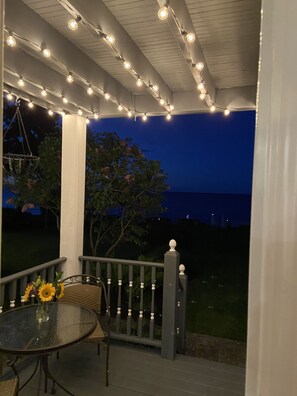 Un espace extérieur aménagé en terrasse, avec des guirlandes lumineuses, une table ornée d’un vase de fleurs et une vue sur l’océan au crépuscule.