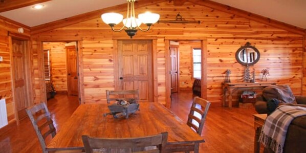 Interior de una cabaña de madera con una mesa de comedor, sillas, una araña y una chimenea.