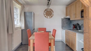 A compact kitchen with wooden cabinets, a dining table with four chairs, a large wall clock, and a modern pendant light. - Bright and spacious apartment close to city center (Châtel)