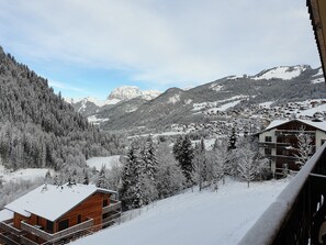 Un chalet de madera con balcón, rodeado de árboles cubiertos de nieve y montañas al fondo.