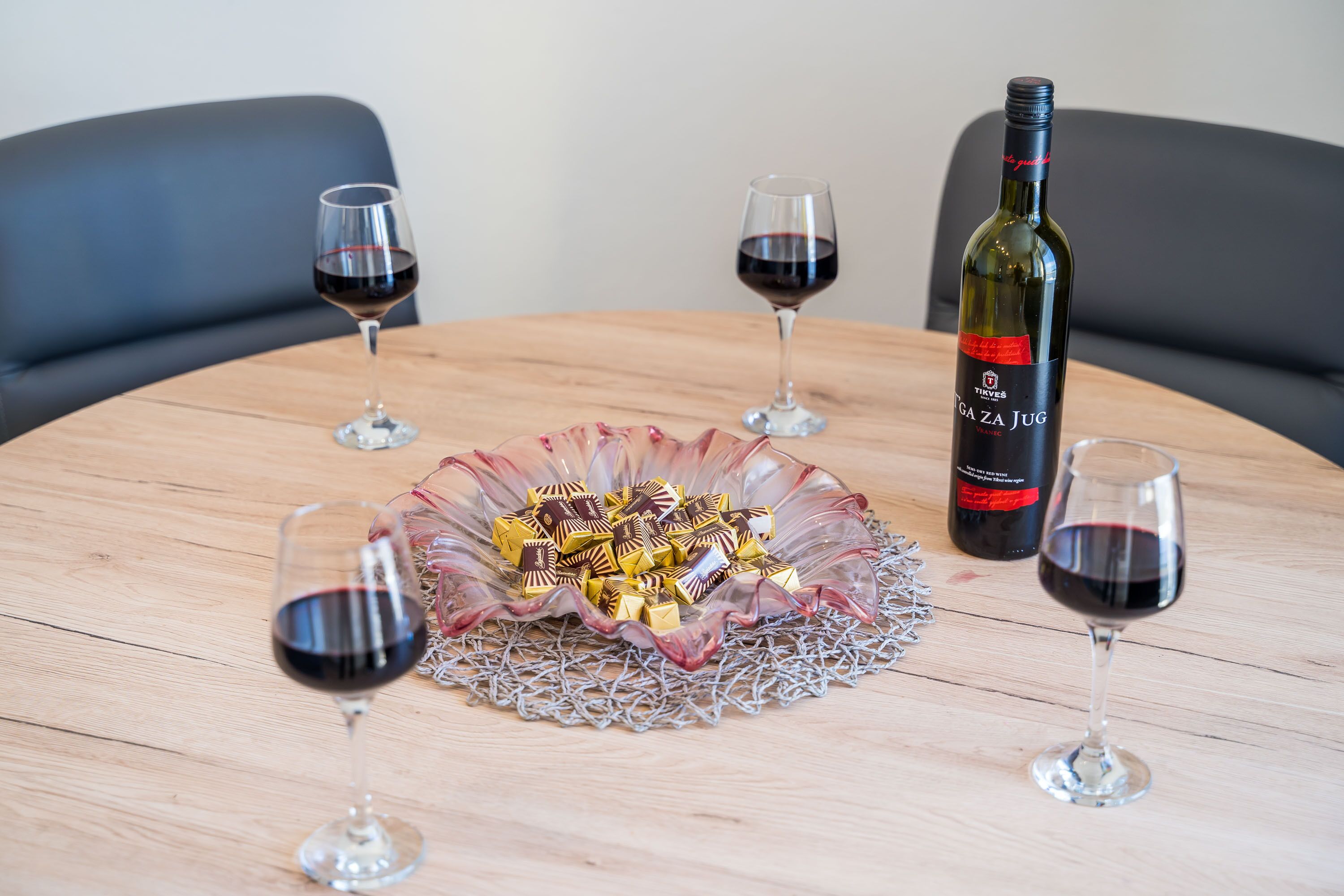 Une table ronde en bois, avec une bouteille de vin, deux verres de vin rouge et un bol de chocolats.