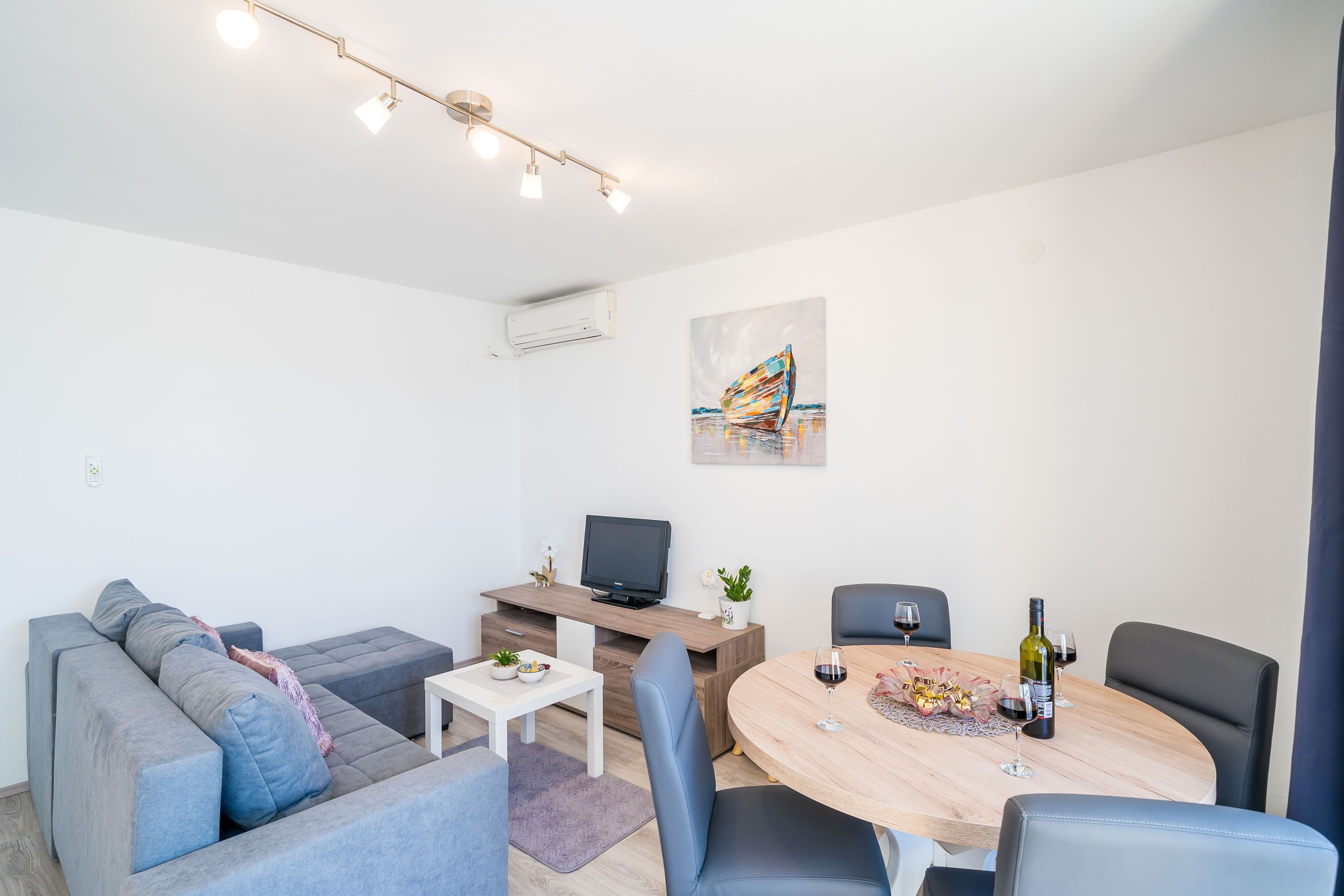 A modern living room with a grey sofa, a small white table, a wooden dining table with chairs, a flat-screen TV on a wooden stand, and a painting on the wall.