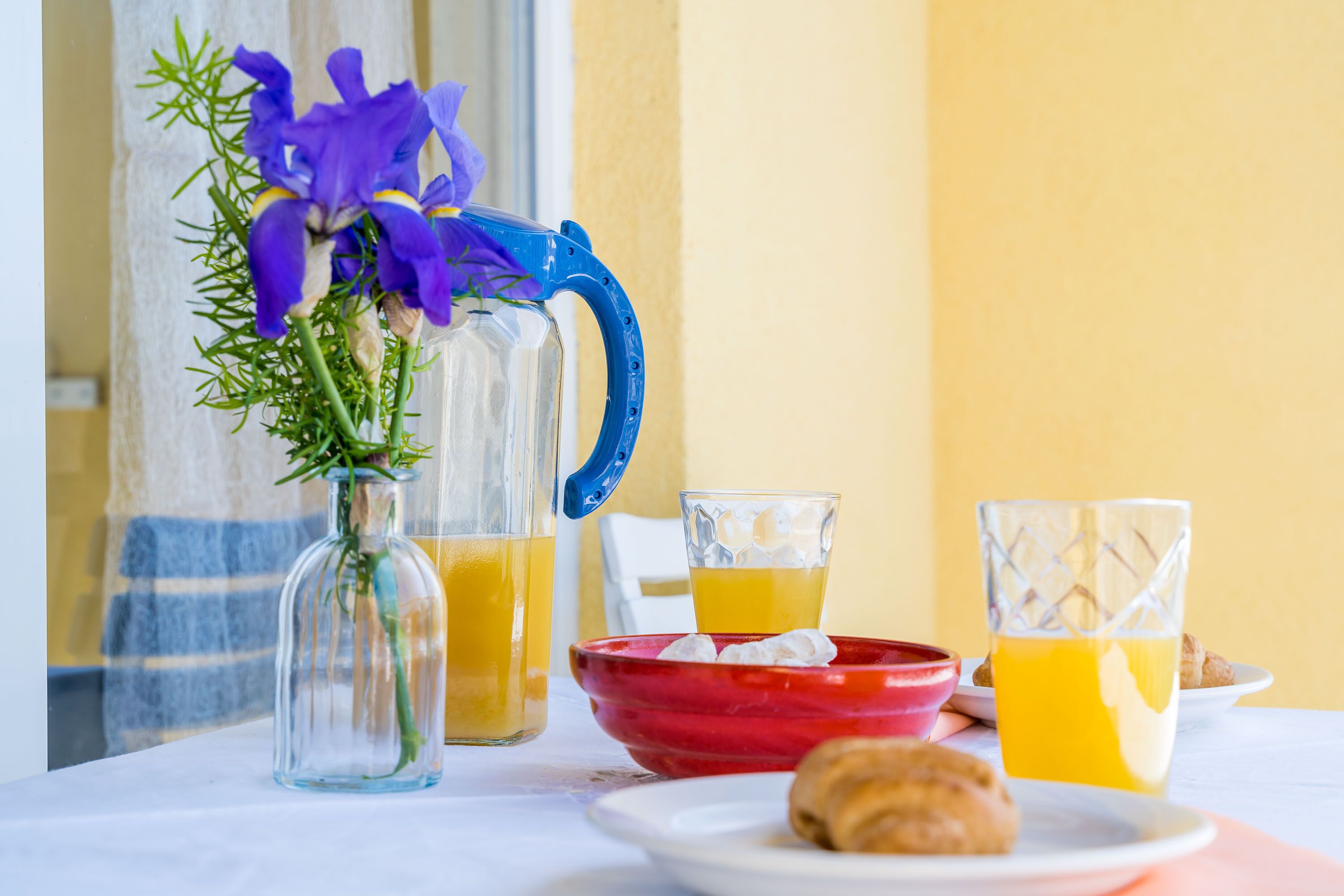 A table with a pitcher of orange juice, a glass of orange juice, a red bowl, and a plate with a pastry.