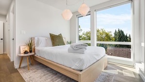 Een moderne slaapkamer met een houten bed, wit beddengoed en een uitzicht op groen door grote ramen.