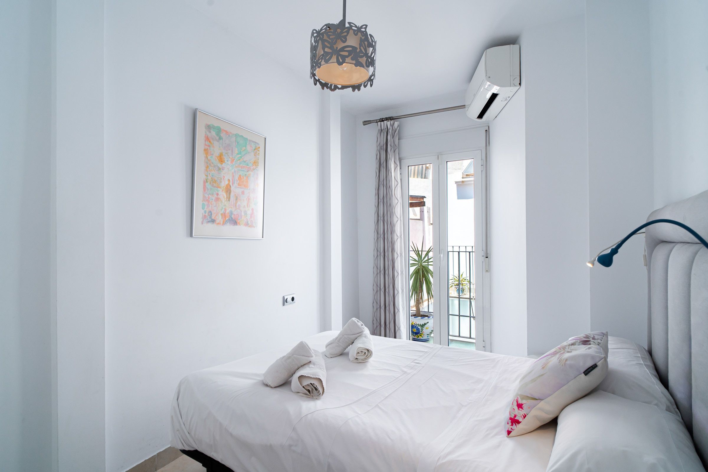 A hotel room with a bed, a hanging light fixture, an air conditioning unit, a door leading outside, and a window with a view of the outside.