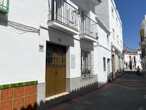 A narrow street with white buildings, a wooden door, and a small balcony with metal railings.