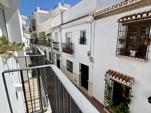 A narrow street with white buildings, balconies with potted plants, and a metal railing.
