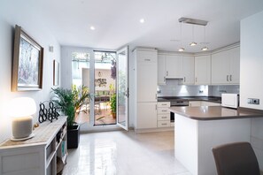 Een moderne keuken met witte kastjes, een eethoek met een tafel en stoelen, en een uitzicht door een glazen deur op een terras.