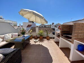 Een terras op het dak met een tafel, stoelen, potplanten, een barbecue en een grote parasol.