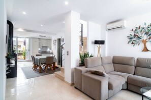 A modern living room with a sofa, a dining area with a table and chairs, and a kitchen in the background.